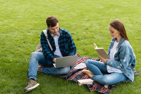 cheerful girl reading book near handsome student using laptop, online study concept
