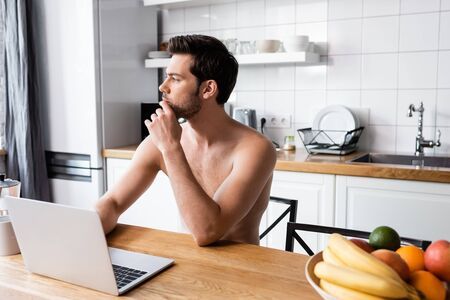 pensive shirtless freelancer working on laptop on kitchenの写真素材