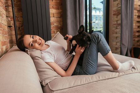happy woman touching cute french bulldog and looking at camera in living roomの写真素材