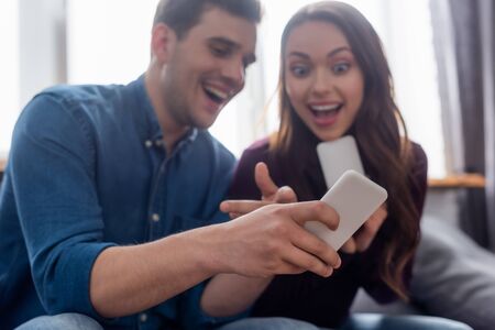selective focus of cheerful man pointing with finger at smartphone near excited girlの写真素材