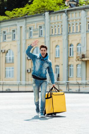 Positive courier with thermo bag waving hand on urban streetの写真素材