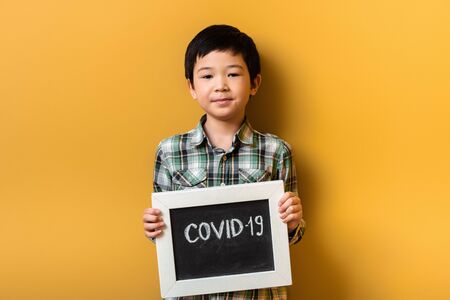 smiling asian boy holding board with covid-19 sign on yellow during quarantineの写真素材
