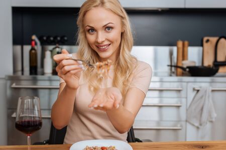 cheerful woman holding fork with thai noodles while smiling at camera near glass of red wineの写真素材