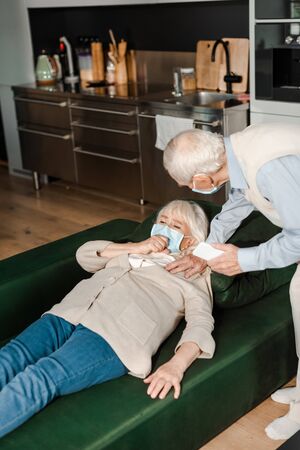 elderly man in medical mask calling doctor with smartphone while ill wife lying on sofa during coronavirus epidemicの写真素材