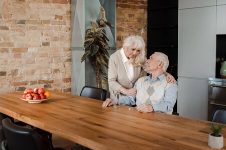 sad elderly wife and sick husband with pills and glass of water at home during quarantine