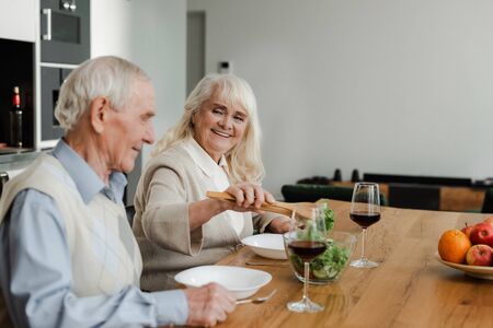 smiling elderly couple having dinner with wine and salad at home on self isolationの写真素材
