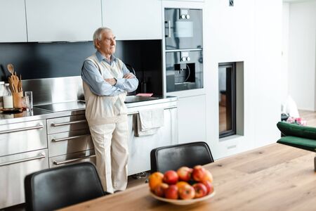 upset elderly man standing in kitchen with fruits on table during quarantineの写真素材