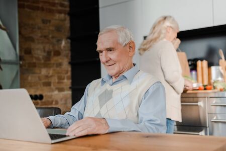 senior wife cooking while happy husband using laptop on kitchen during self isolationの写真素材