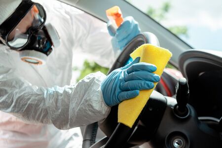 specialist in hazmat suit and respirator cleaning car interior with antiseptic spray and rag during...