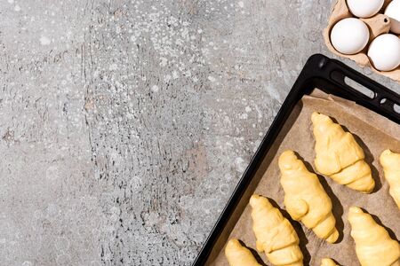 top view of uncooked croissants on baking tray near eggs on concrete grey surfaceの写真素材