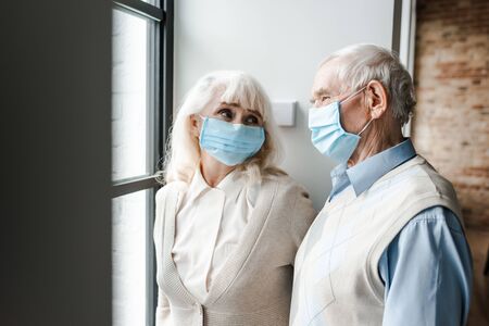 elderly couple in medical masks looking through window during quarantine