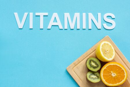 top view of ripe orange, lemon and kiwi halves on wooden cutting board and word vitamins on blue...