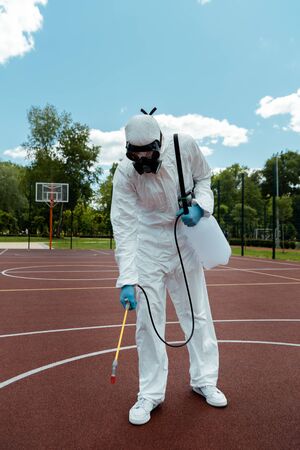 worker in hazmat suit and respirator disinfecting basketball court in park during covid-19 pandemic