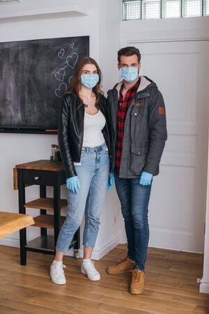 young couple in protective masks and latex gloves looking at camera while standing near door