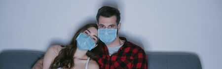 horizontal image of young couple in medical masks watching tv on sofa at homeの写真素材