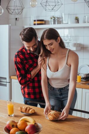 handsome man touching shoulder of pretty girlfriend holding plate with tasty croissant near fresh fruitsの写真素材