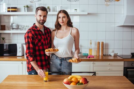 smiling girl holding plates with delicious croissant near boyfriend touching glass of orange juiceの写真素材
