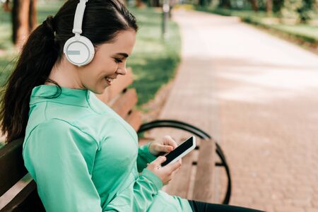 Side view of smiling sportswoman in headphones using smartphone on bench in parkの写真素材