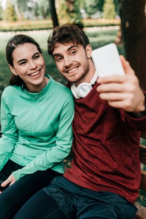 Selective focus of smiling sportsman taking selfie with smartphone near girlfriend on bench in parkの写真素材