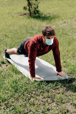 Sportsman in medical mask doing press ups while training on fitness mat in park