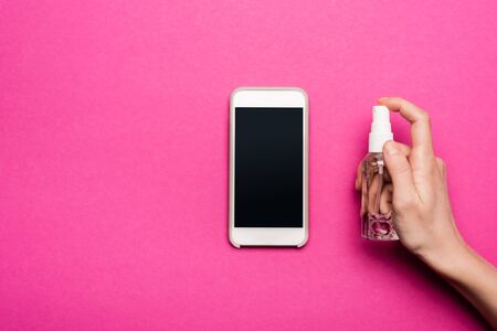 partial view of woman spraying antiseptic on smartphone with blank screen on pinkの写真素材