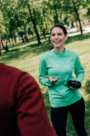 Selective focus of smiling sportswoman talking to boyfriend while holding sports bottle in parkの写真素材