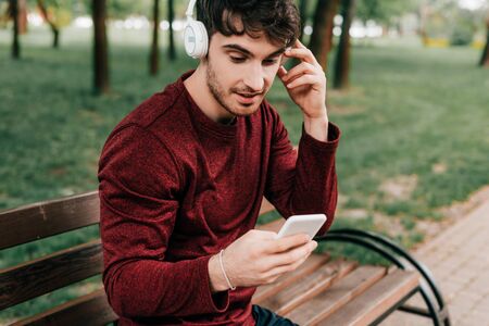 Handsome sportsman listening music in headphones and using smartphone on bench in parkの写真素材