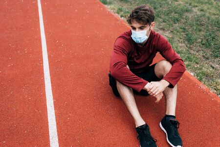 Sportsman in medical mask sitting on running track in parkの写真素材