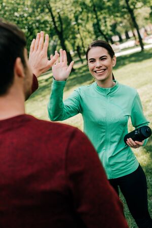 Selective focus of smiling sportswoman high five to man while holding sports bottle in park