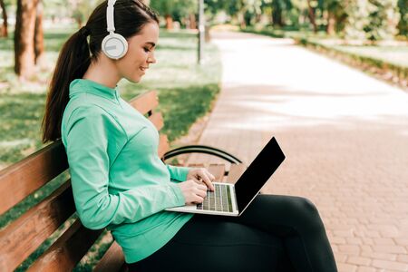 Side view of attractive smiling freelancer in headphones using laptop on bench in parkの写真素材