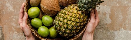 cropped view of woman holding ripe exotic fruits in wicker basket on weathered surface, panoramic cropの写真素材