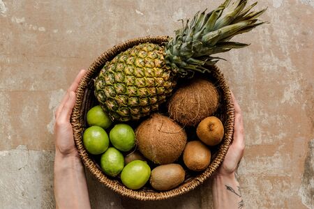 cropped view of woman holding ripe exotic fruits in wicker basket on weathered surfaceの写真素材