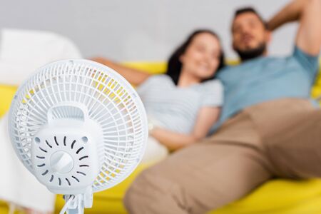 Selective focus of electric fan near young couple sitting on sofaの写真素材