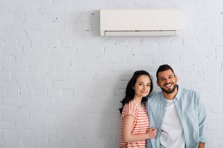 Young couple smiling at camera while standing near air conditioner on wall at homeの写真素材