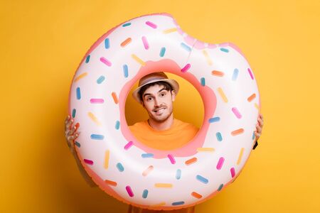 cheerful man in hat standing with inflatable donut on yellow