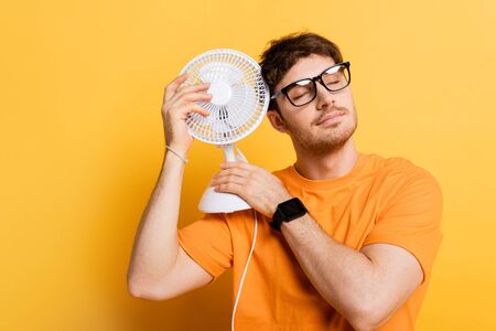 dreamy man with closed eyes in eyeglasses holding electric fan on yellow