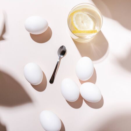 top view of white chicken eggs, water with lemon and teaspoon on grey table with shadows