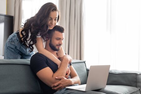 happy girl hugging handsome boyfriend using laptop in living room
