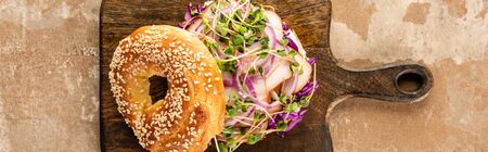 top view of fresh delicious bagel with meat, red onion and sprouts on wooden cutting board on aged beige surface, panoramic shotの写真素材