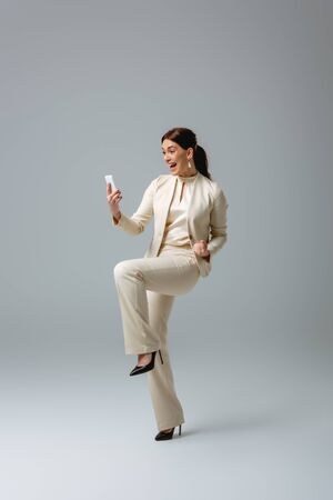 Cheerful woman in formal wear showing yes gesture while using smartphone on grey backgroundの写真素材