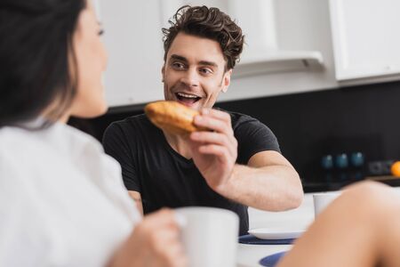 Selective focus of positive man holding croissant near girlfriend with cup of coffeeの写真素材