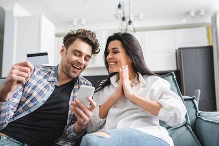 Selective focus of smiling man holding credit card and smartphone near girlfriend showing please gesture at homeの写真素材