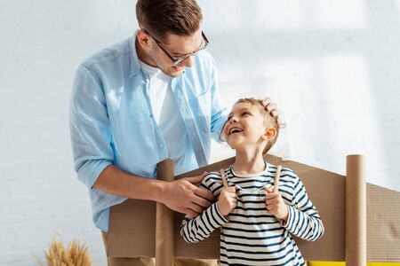 happy father touching head of cheerful son with cardboard plane wings on backの写真素材