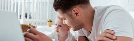 panoramic shot of young father and cute little son on bed near laptop