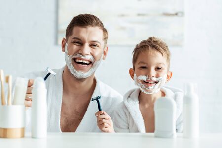 selective focus of cheerful father and son with shaving foam on faces holding shaving razorsの写真素材