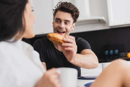 Selective focus of positive man holding croissant near girlfriend with cup of coffeeの写真素材