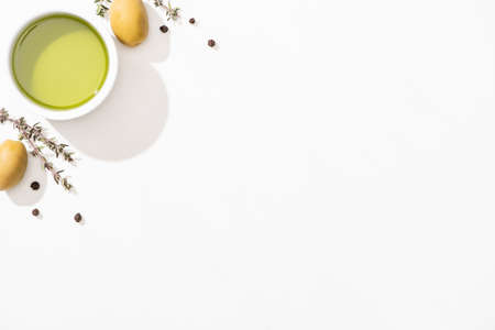 top view of olive oil in bowl near green olives, herb and black pepper on white background