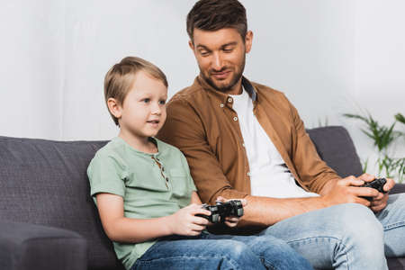 KYIV, UKRAINE - JUNE 9, 2020: happy father looking at smiling son while playing video game with...