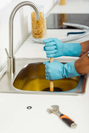 Cropped view of workman in rubber gloves cleaning kitchen sink with plunger near wrench on worktopの写真素材
