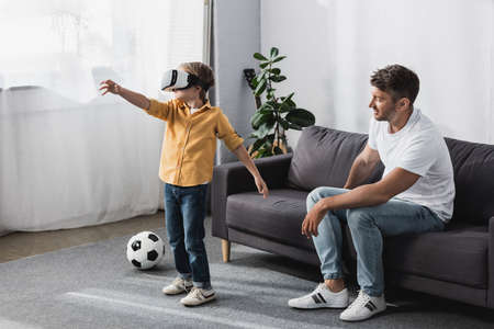 boy in vr headset standing with outstretched hand near father sitting on sofa